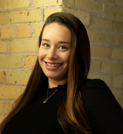 Woman with long brown hair smiles, standing by a brick wall.
