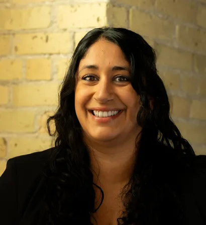 Smiling woman with dark curly hair against a brick wall.