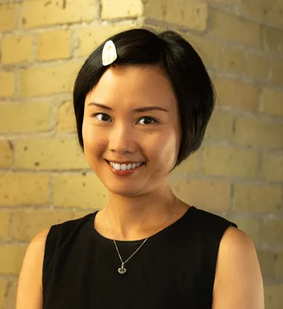 Smiling woman with short dark hair and a white hair clip, posing in front of a brick wall.