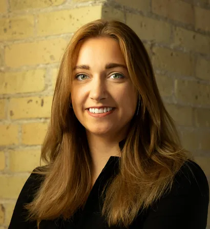 Smiling woman with long blonde hair, wearing a black shirt, and standing near a brick wall.