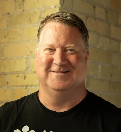 Man smiling, standing in front of a brick wall.