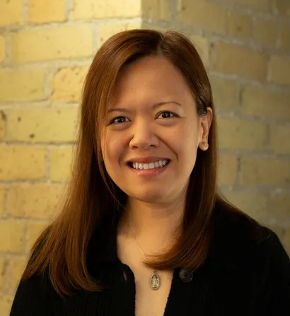 Smiling woman with long brown hair, wearing a black shirt, beside a brick wall.